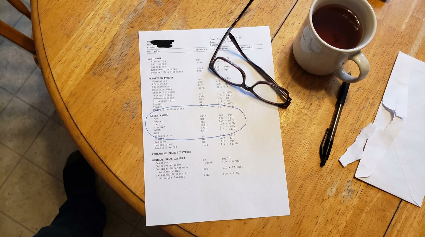 A printed blood test result on a kitchen table, reading glasses and a mug beside it, the liver panel circled in biro
